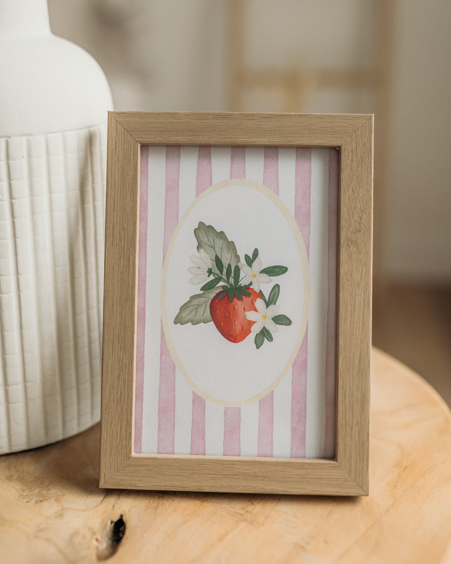 Framed artwork with a strawberry illustration on a striped background, placed on a wooden surface.